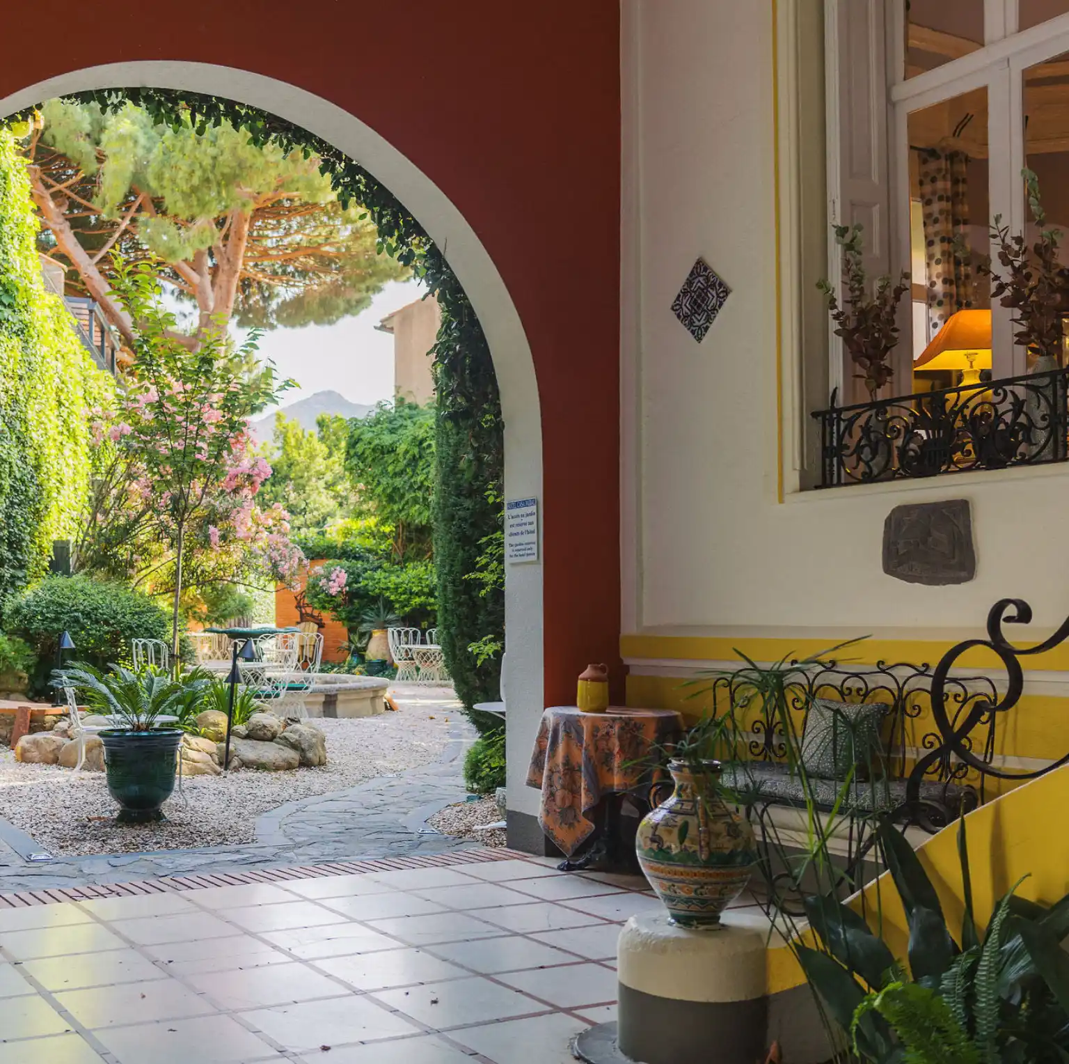 A cozy outdoor garden patio with a pathway leading through a lush courtyard featuring green plants, blooming pink flowers, and white wrought iron chairs and tables surrounding a central stone fountain, viewed through a white and red archway connected to a yellow and white tiled porch with decorative vases and a wrought iron bench accented with cushions.