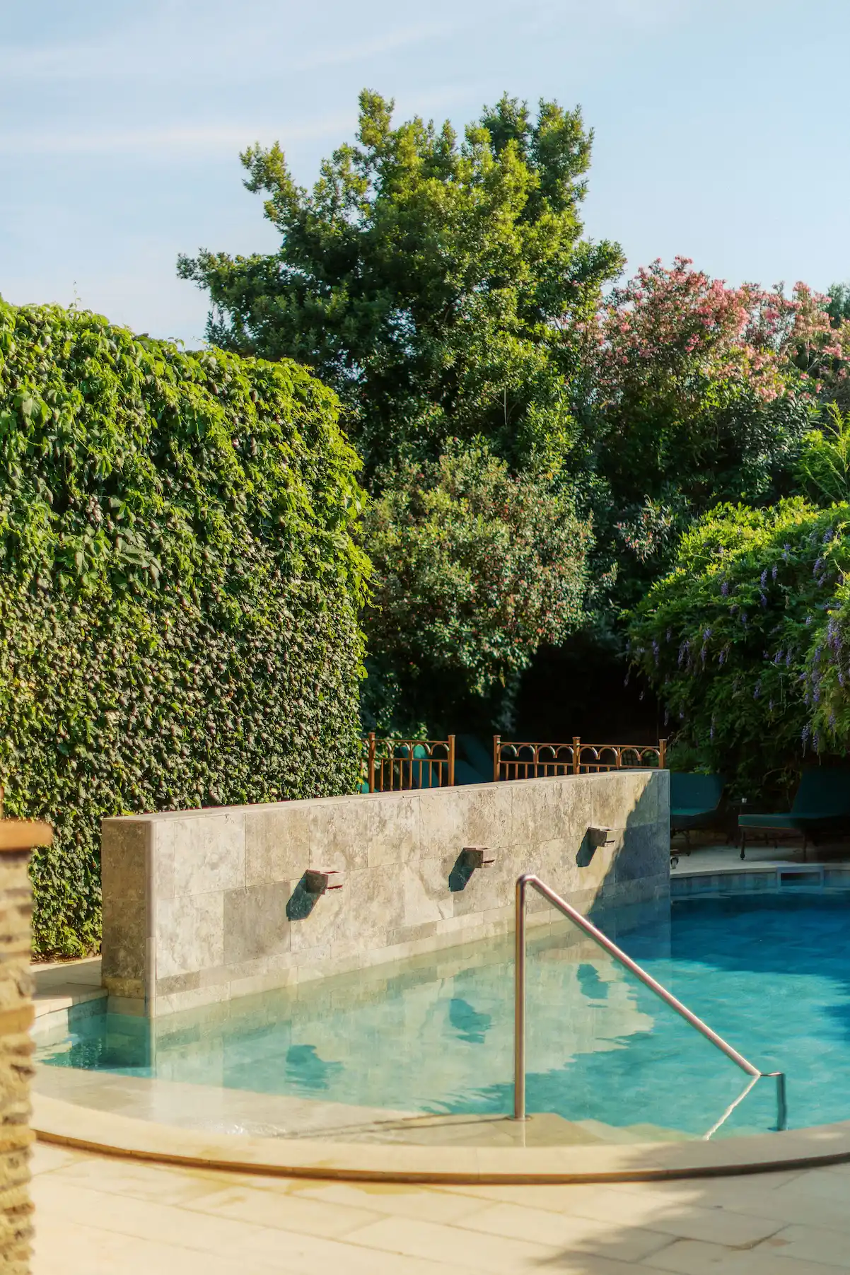Outdoor swimming pool with clear blue water, a stone wall with three water spouts, metal handrail, and lush green trees and shrubs surrounding the area.