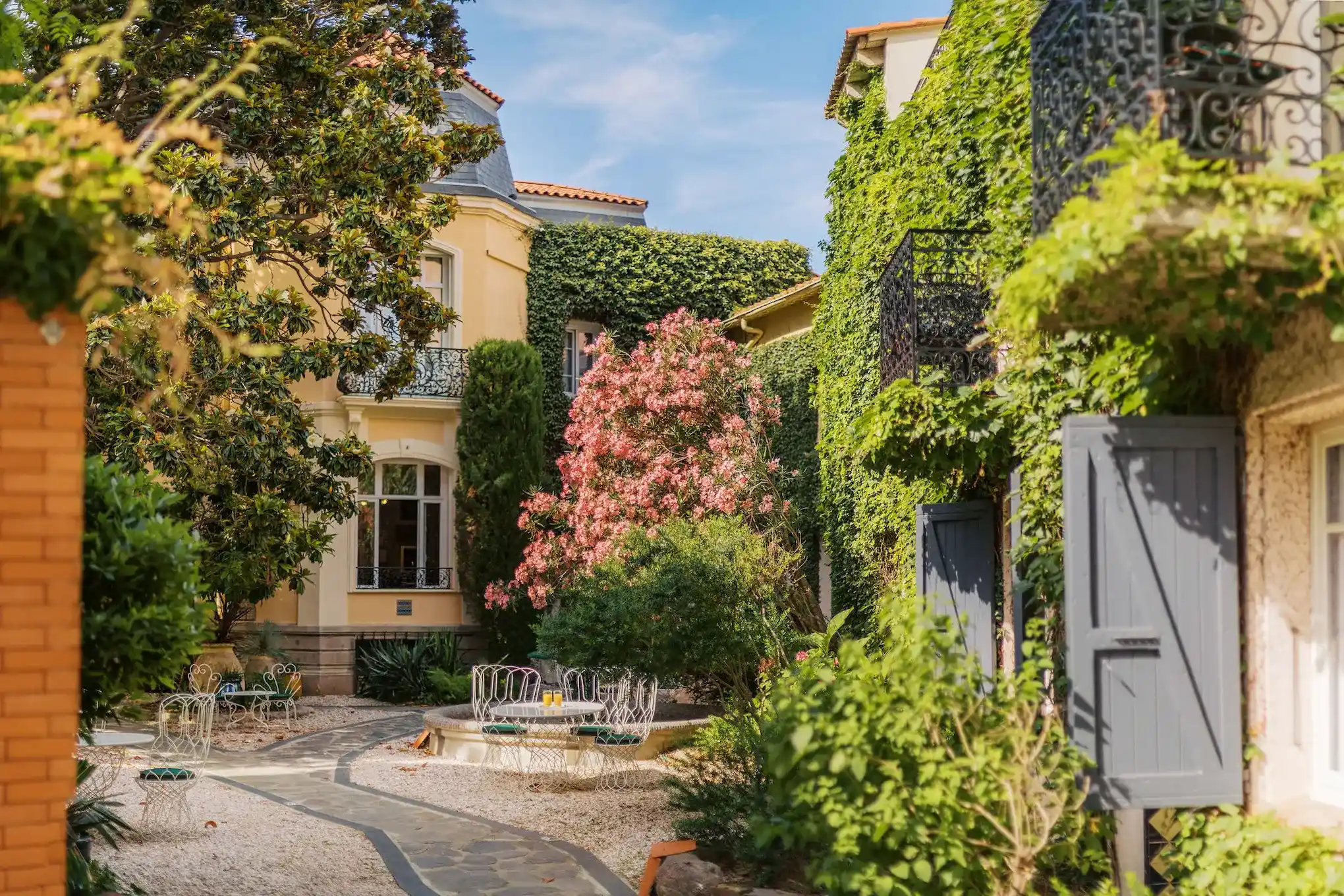 Sunny courtyard with lush greenery and blooming pink flowers surrounded by ivy-covered buildings, featuring a stone pathway and white wrought iron chairs with green cushions around a circular table.