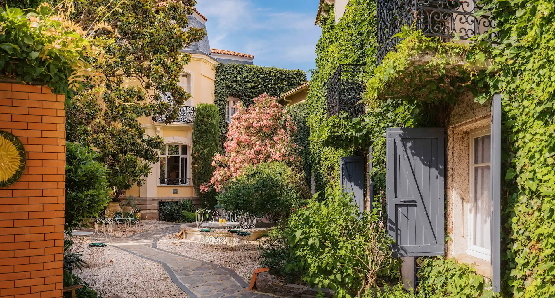 Sunlit courtyard garden with a stone path, ivy-covered walls, open gray shutters on windows, pink flowering bush, green trees, and white metal chairs around tables.