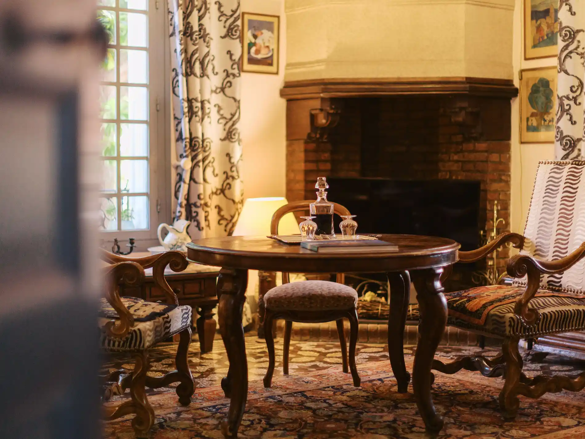 Cozy living room with a round wooden table surrounded by ornate wooden chairs with patterned upholstery, a brick fireplace with a wooden mantel in the background, and a window with patterned curtains letting in natural light.