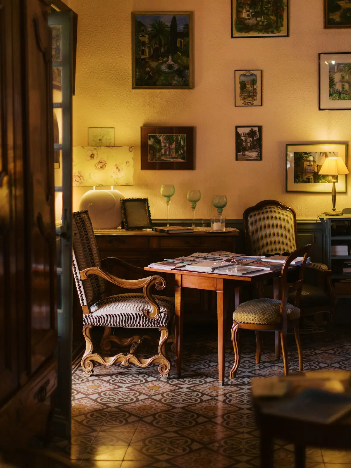 Cozy room with antique wooden chairs and a table covered in magazines and papers, illuminated by warm lamps, with framed paintings on the textured yellow walls and decorative patterned floor tiles.