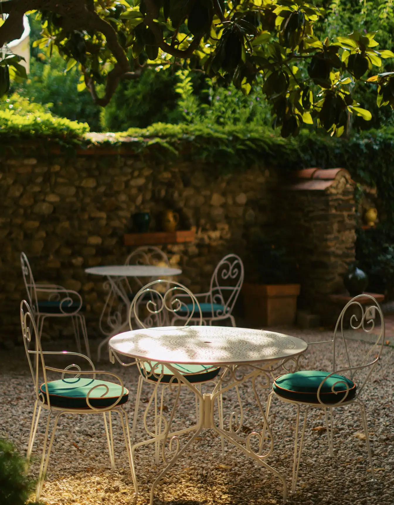 Outdoor patio area with vintage white wrought iron tables and chairs with green cushions, surrounded by gravel ground, a stone wall, and leafy greenery casting shadows.