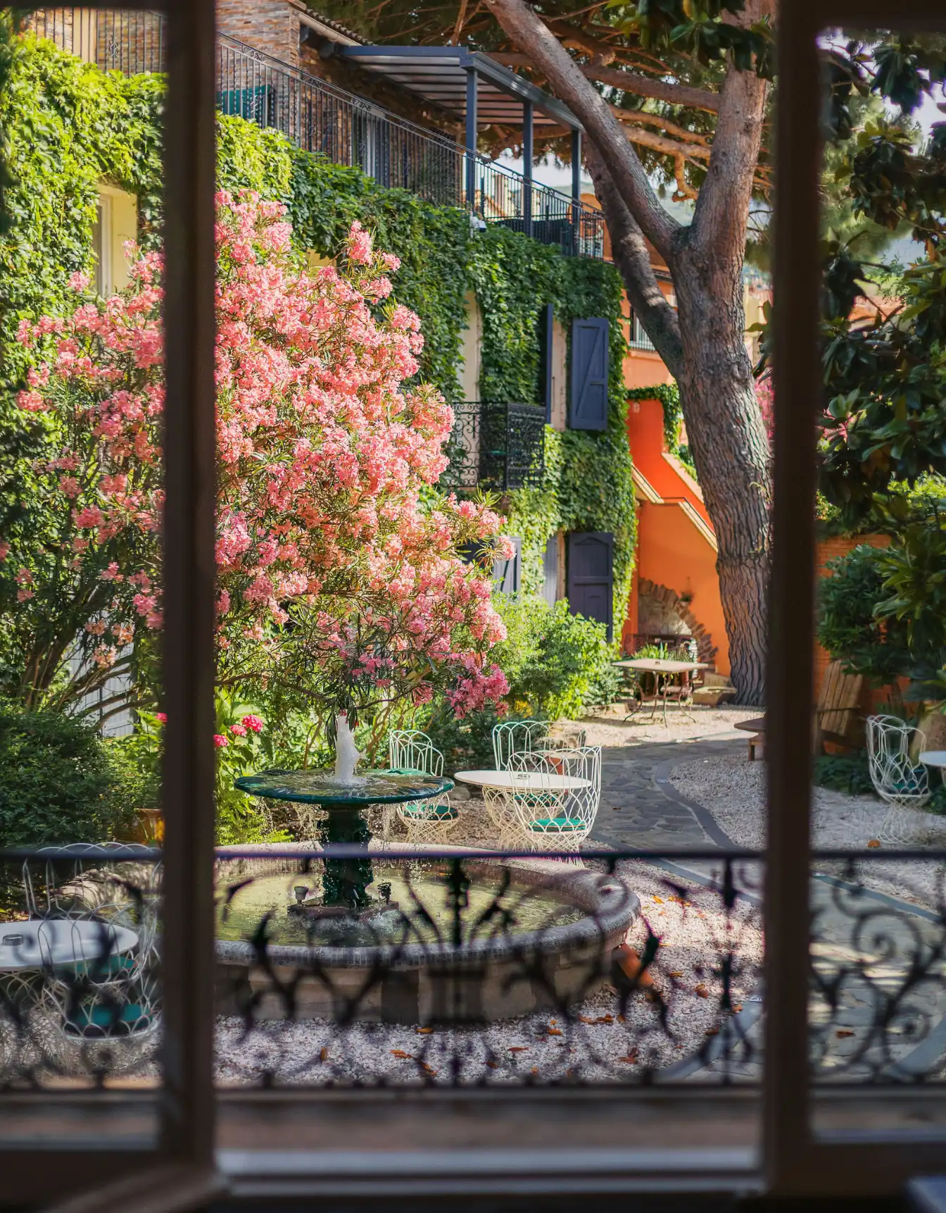 View through window of a courtyard with a central water fountain surrounded by white metal chairs and tables, flowering pink bushes, and a building covered with green ivy and blue shutters.