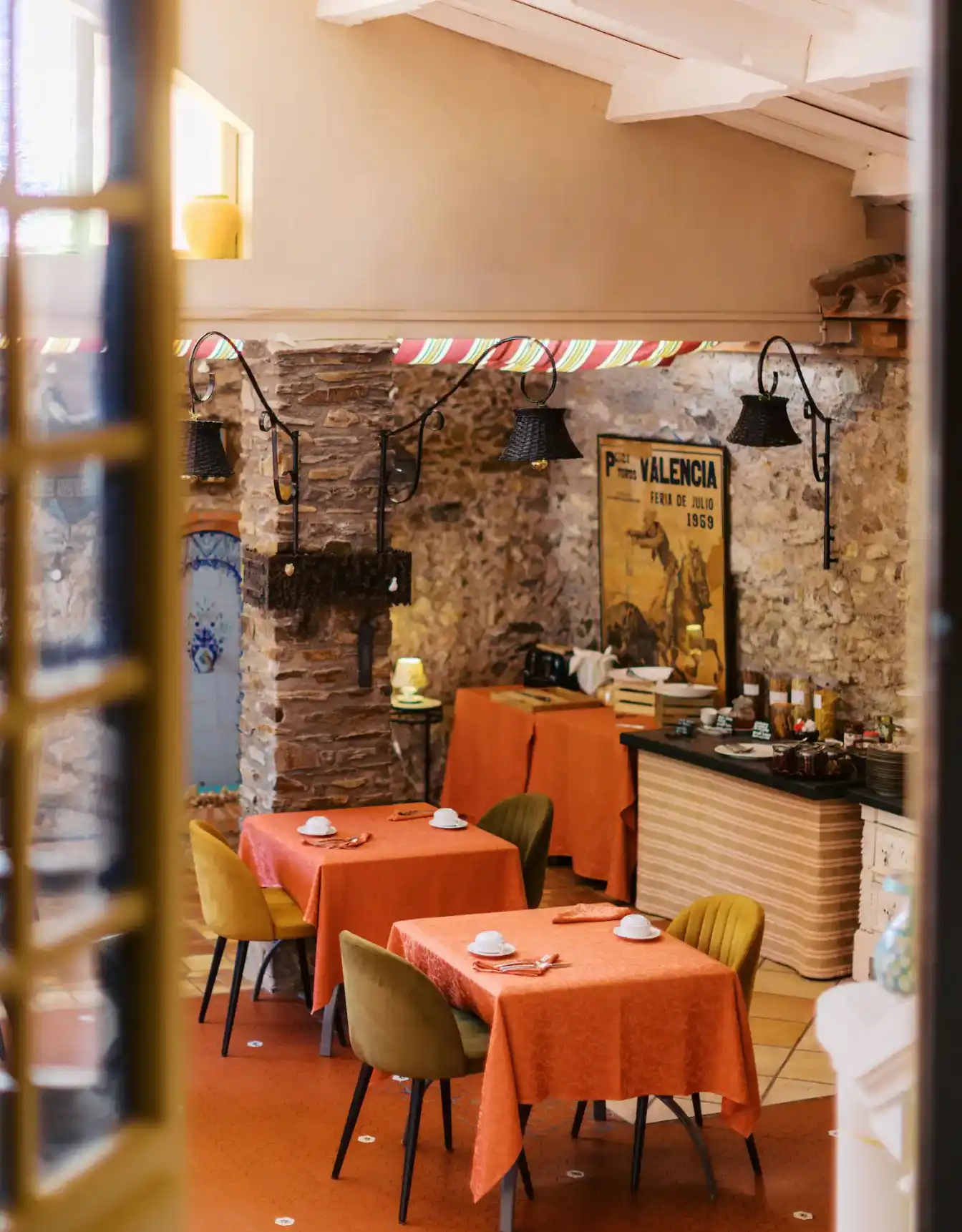 Cozy dining area with two tables covered in orange tablecloths, each set with white cups and silverware, surrounded by green and yellow chairs; stone walls, vintage wall lamps, and a large poster reading 'Valencia Feria de Julio 1969' decorate the space.