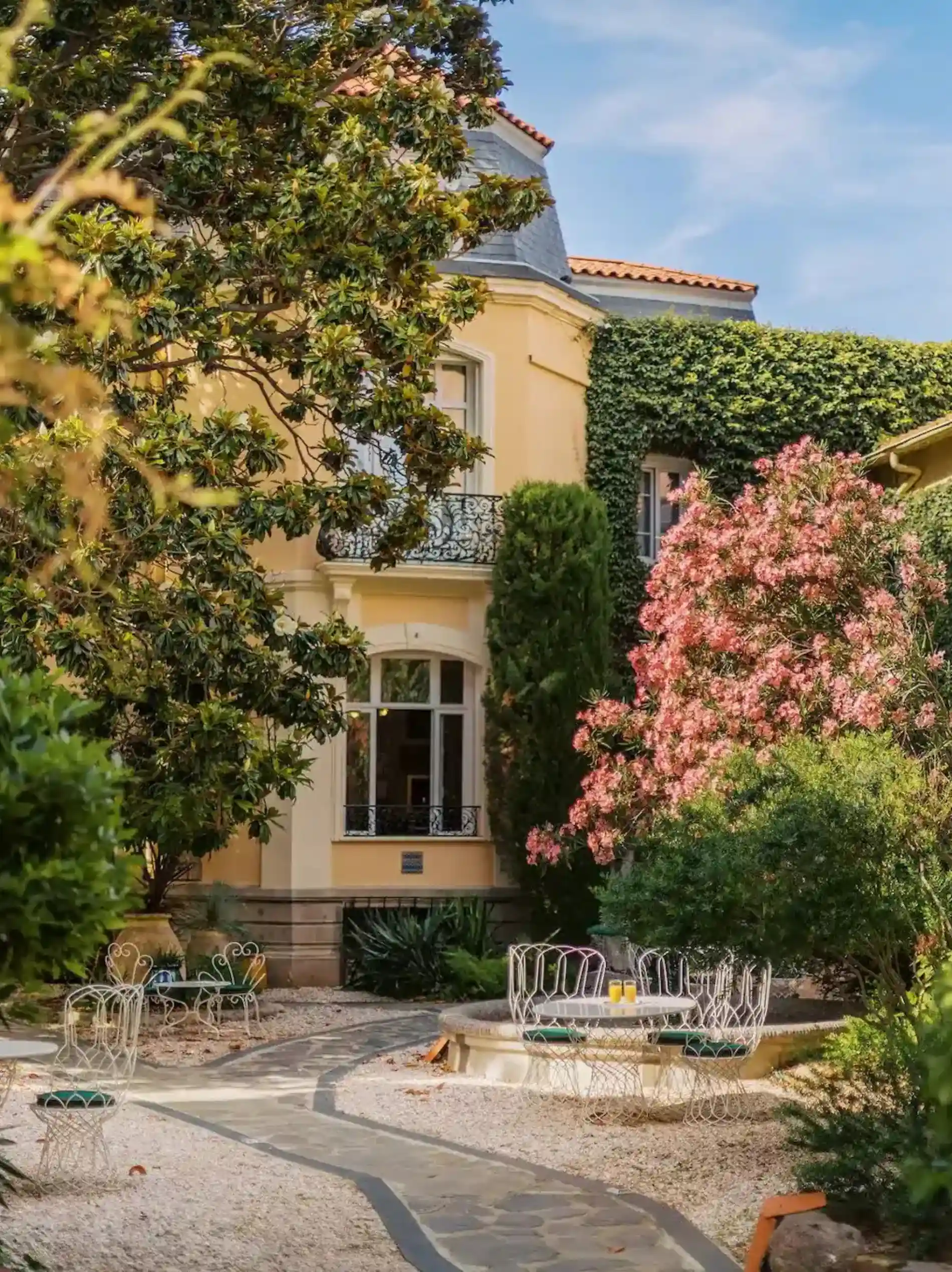 Outdoor garden area with a stone pathway leading to a round stone table surrounded by white wrought iron chairs with green cushions, in front of a yellow house partially covered in ivy, with trees and pink and green flowering bushes around.