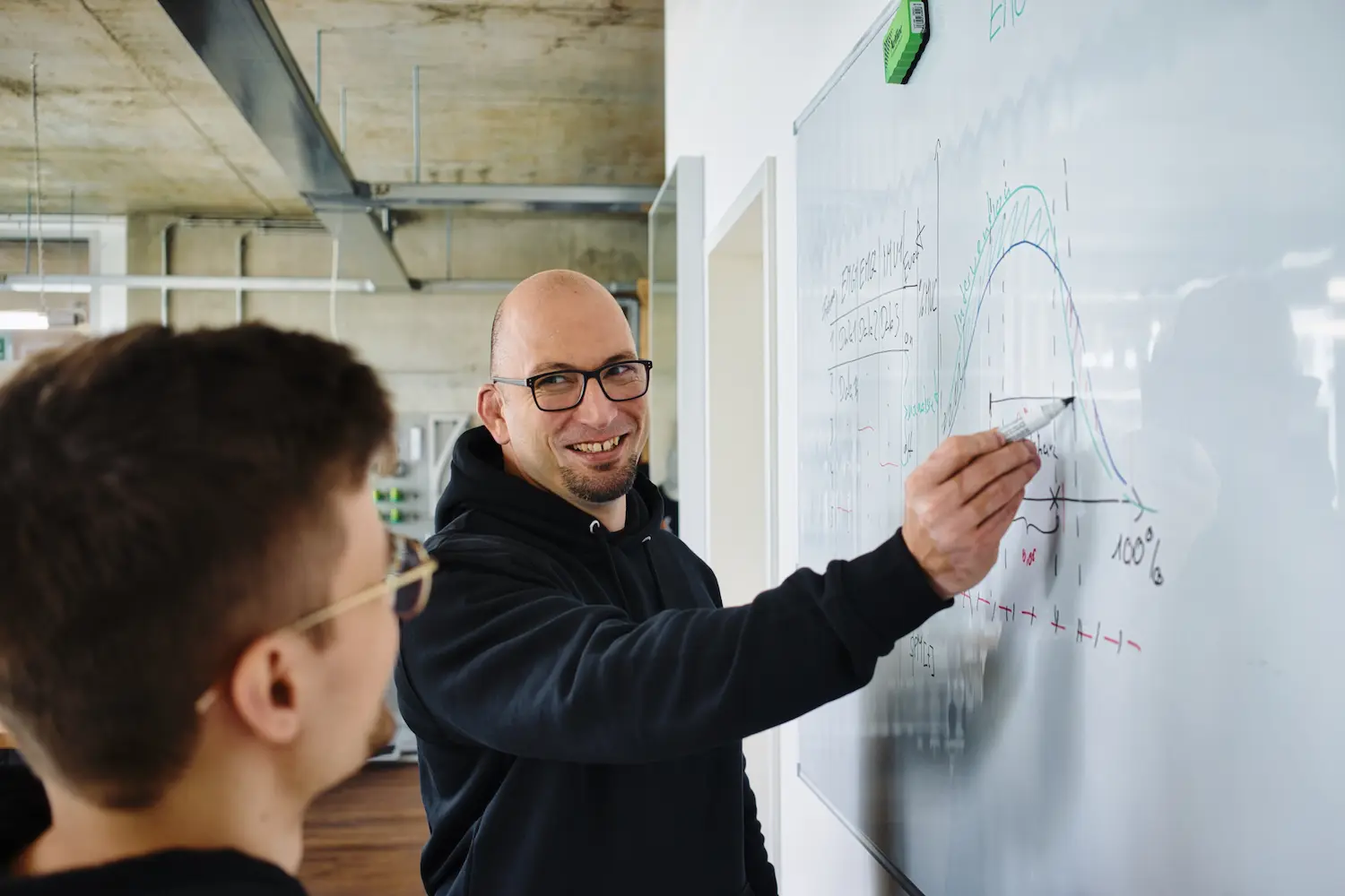 A man in a black hoodie points at a white board with a graph on it.