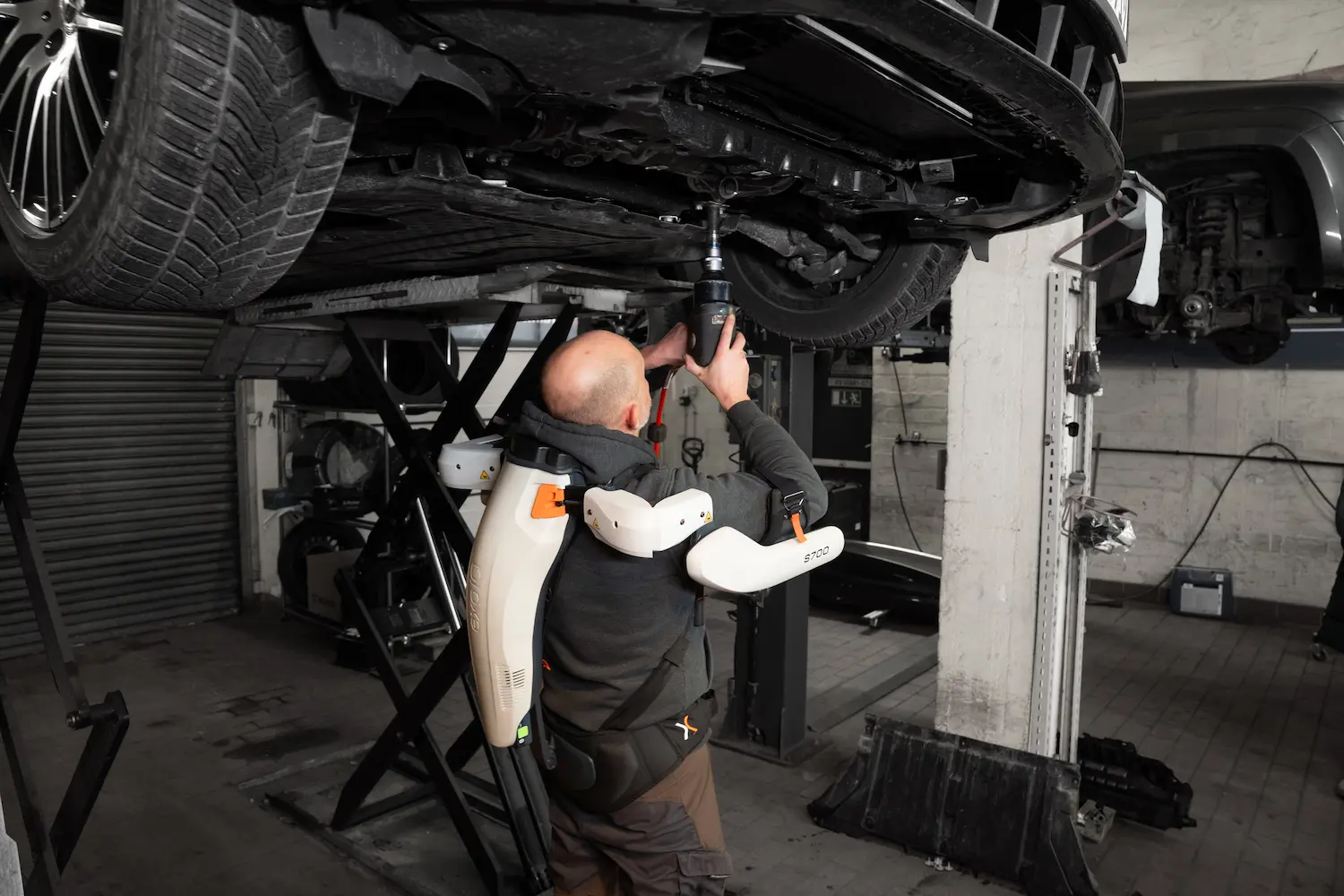 A man is working on a car in a garage with an exoskeleton.