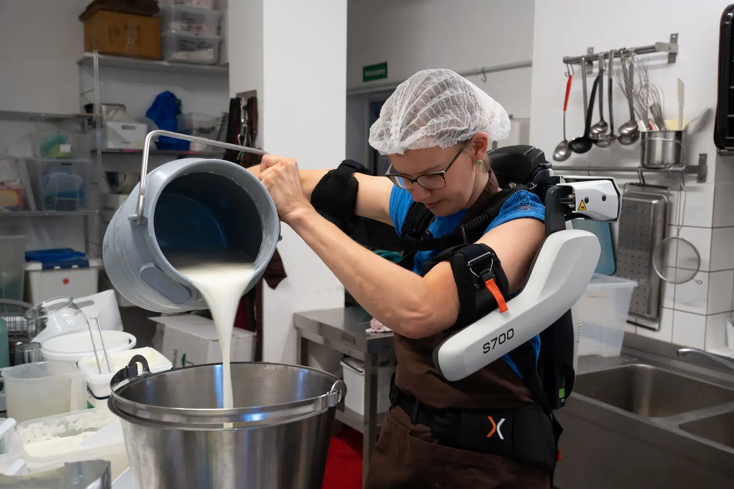 A woman wearing a white plastic bag on her head pouring milk into a bucket with an exoskeleton.