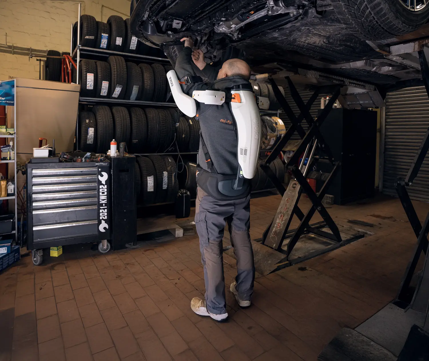 A man is working on a car in a garage with an exoskeleton.