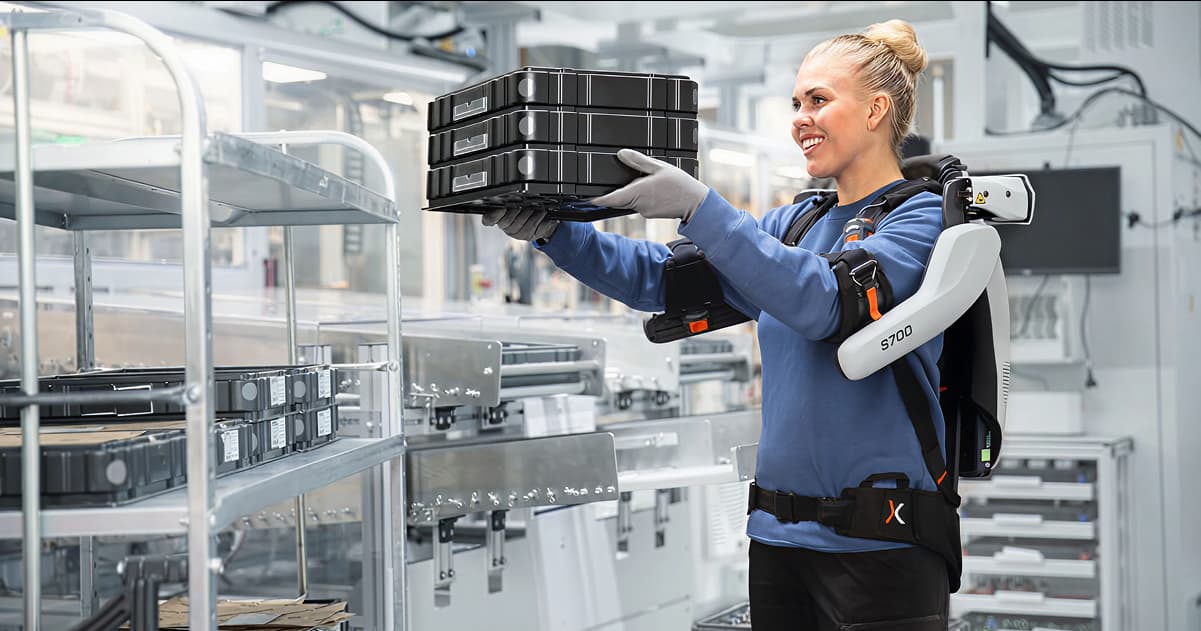 A woman wearing a blue shirt and black gloves is holding a stack of black boxes with an exoskeleton.