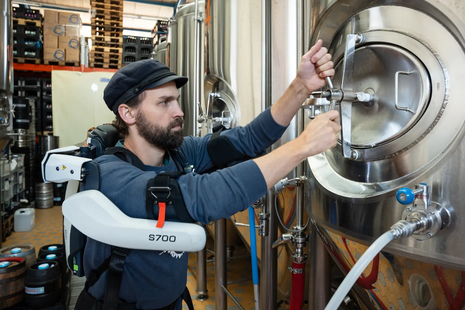 A man wearing a exoskeleton and a hat is adjusting a valve on a large metal tank.