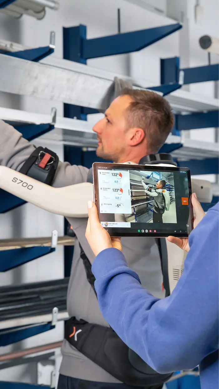 The exoIQ app on a tablet in the foreground, a person wearing an active S700 shoulder exoskeleton in the background