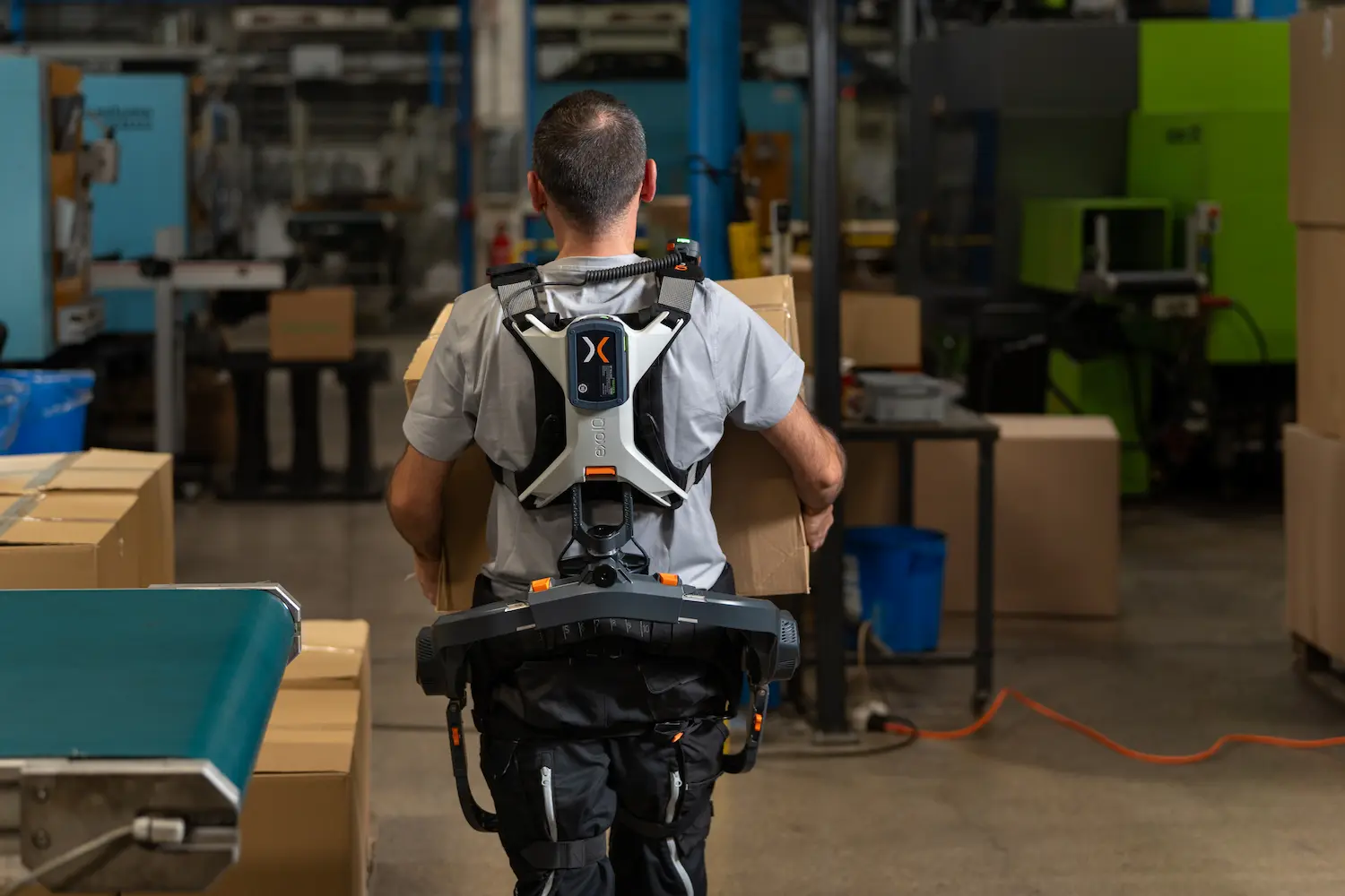 Ein Mann mit Rücken-Exoskelett trägt einen Karton durch eine Fabrikhalle