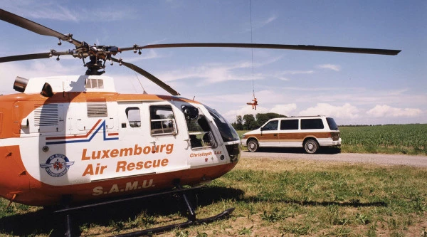 Hélicoptère de Luxembourg Air Rescue stationné dans un champ avec un véhicule à proximité et une opération de treuillage en cours en arrière-plan.