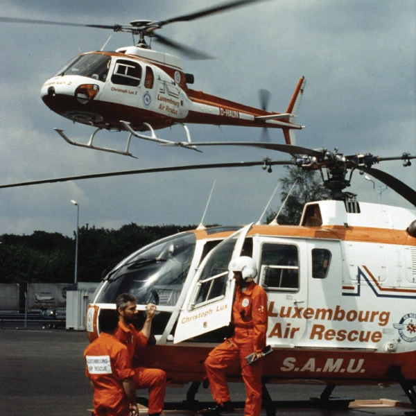Deux membres d’équipage en combinaison orange près d’un hélicoptère Luxembourg Air Rescue, avec un autre hélicoptère en vol au-dessus.