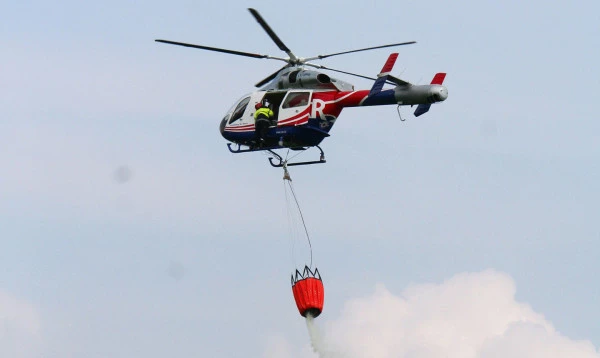 Hélicoptère Luxembourg Air Rescue effectuant une opération de lutte contre les incendies avec un seau d’eau.