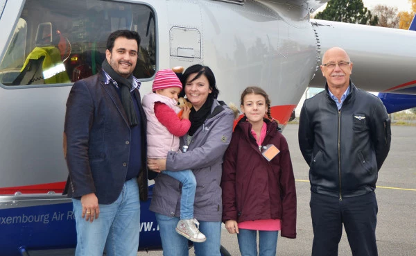 Équipage Luxembourg Air Rescue avec une famille et des enfants secourus devant un hélicoptère, illustrant les soins et l’aide humanitaire.
