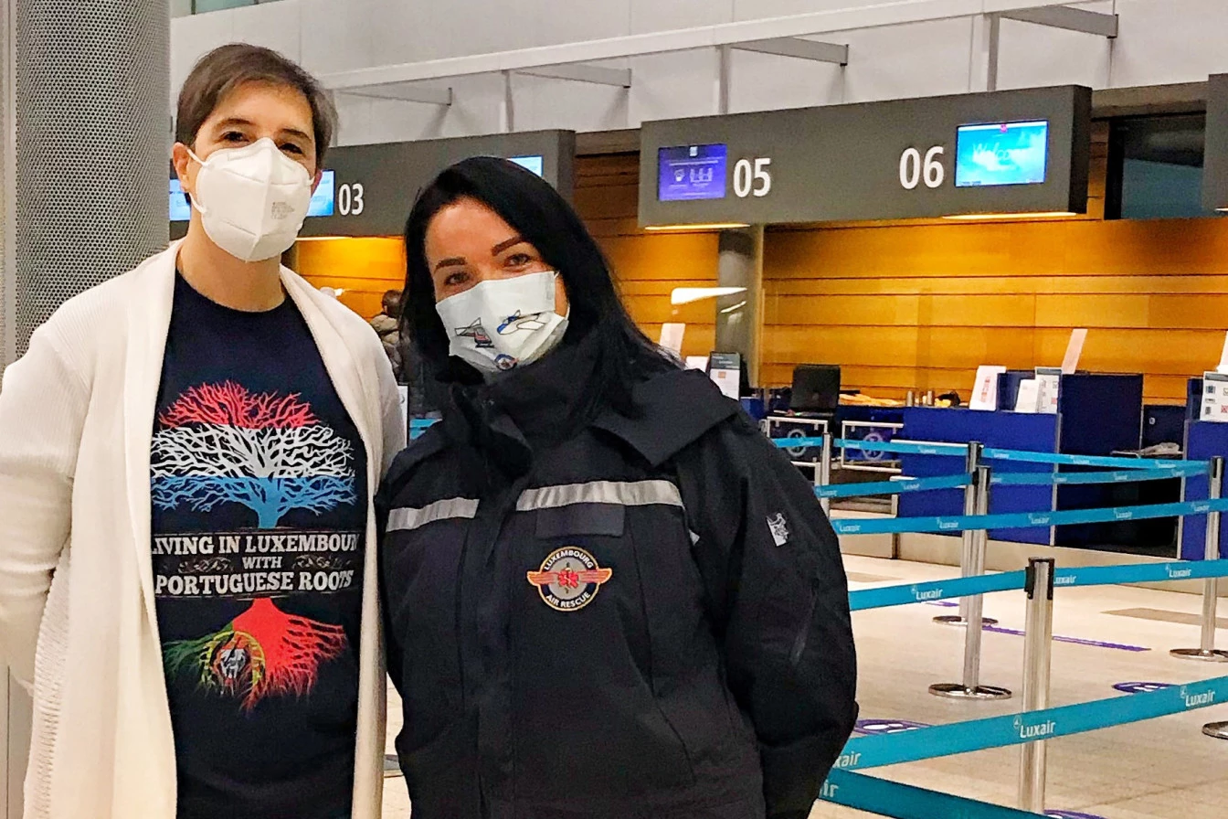 Two women wearing face masks, including a Luxembourg Air Rescue member, standing at an airport check-in area in front of Luxair counters.