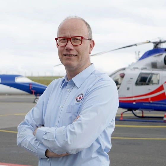 Luxembourg Air Rescue staff member with red glasses standing with arms crossed in front of a helicopter and aircraft on the tarmac.