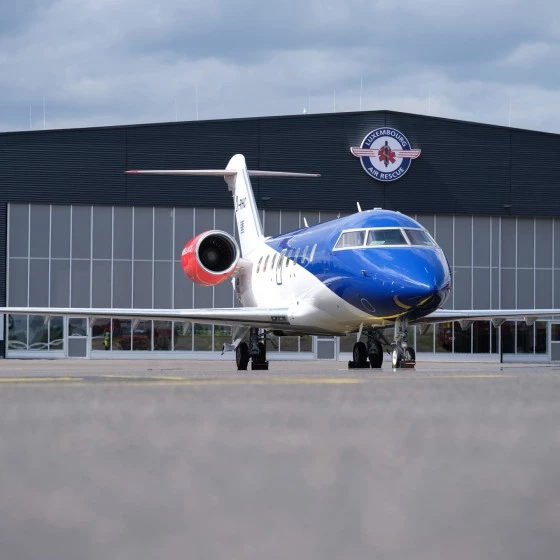 Front view of a small private jet parked on an airport tarmac, with a blue nose and red engine accents. The aircraft is positioned in front of a large hangar displaying the Luxembourg Air Rescue logo. The sky is overcast, and the scene is viewed from a low angle emphasizing the plane’s size.