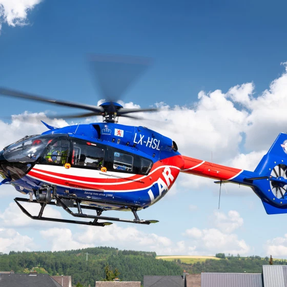 Luxembourg Air Rescue helicopter flying in mid-air against a blue sky with scattered clouds. The aircraft is painted in blue, red, and white, with visible registration markings and a spinning rotor. Below, rooftops and a green landscape can be seen.