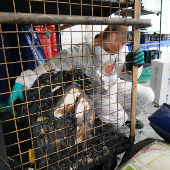A technician wearing protective coveralls, gloves, and a face mask examines a burnt and damaged object inside a metal cage in a workshop. Another person in similar protective gear works in the background among equipment and tools.