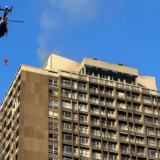 A rescue helicopter hovers above a tall residential building while a person is being lowered on a winch. Light smoke rises from the rooftop against a clear blue sky, suggesting an emergency response.