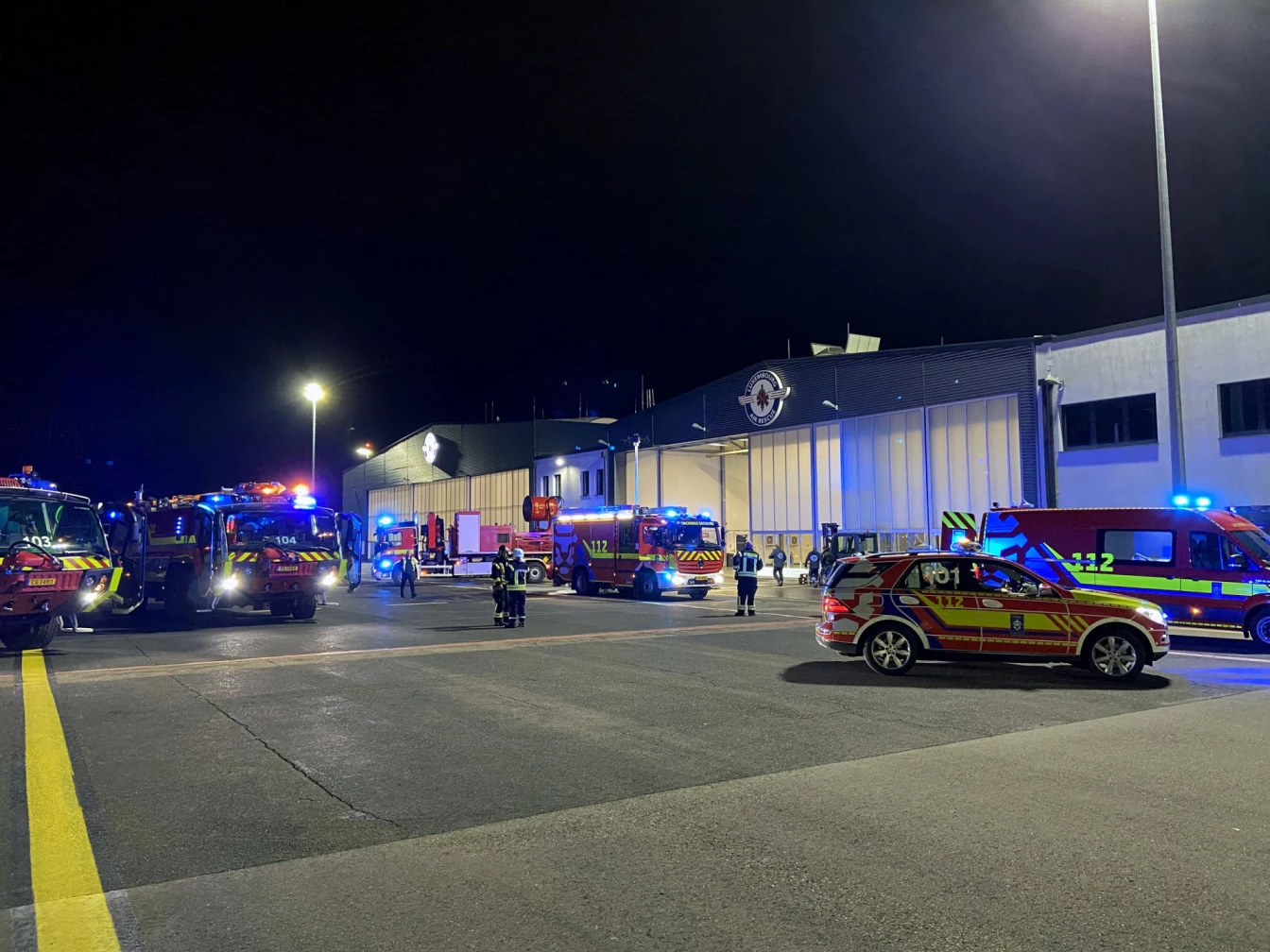 Nighttime emergency scene outside a large hangar, with multiple fire trucks, ambulances, and emergency vehicles with flashing blue lights. Firefighters and responders stand and move around the area, coordinating operations.