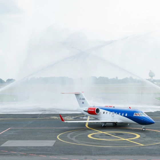 A private jet parked on an airport apron is being sprayed with water from two fire trucks on either side, creating a ceremonial water arch. The scene takes place on a runway under an overcast sky.