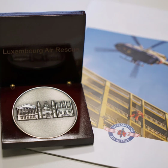 A commemorative medal displayed in an open wooden case labeled “Luxembourg Air Rescue,” featuring an engraved architectural scene. In the background, a brochure shows a rescue helicopter flying near a building.