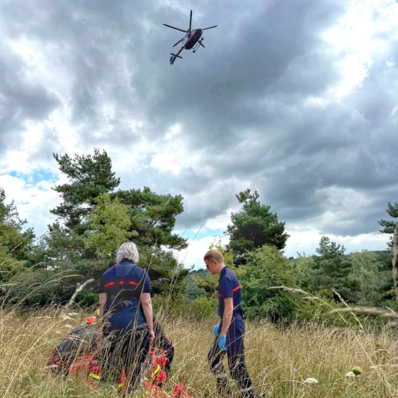 Two rescue workers stand in a grassy field beside a stretcher while a helicopter hovers overhead under a cloudy sky. The scene suggests an ongoing air rescue operation in a natural environment.