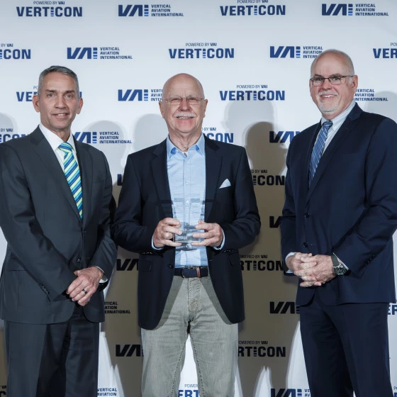 Three men in business attire pose for a photo at an event in front of a backdrop displaying “Vertical Aviation International” and “VERTICON.” The man in the center holds a glass award, suggesting a recognition or ceremony.