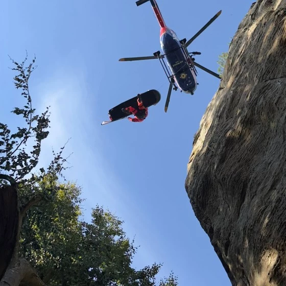 A rescue helicopter hovers above a rocky cliff while a rescuer is lowered on a stretcher by a winch. The scene is viewed from below, with trees and rock formations framing the clear sky.