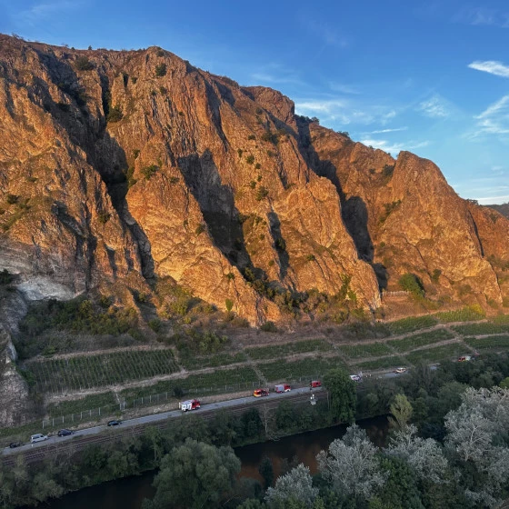 Aerial view of a steep rocky cliff illuminated by warm light, with several emergency vehicles lined up along a road below beside a river. Trees and vegetation surround the area under a clear sky.