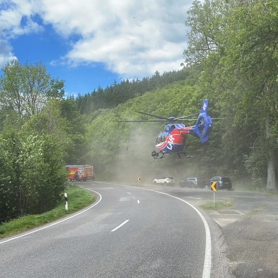 A rescue helicopter hovers low over a curved road in a forested area, raising dust as emergency vehicles and cars are stopped nearby. The scene takes place under a partly cloudy sky surrounded by trees.