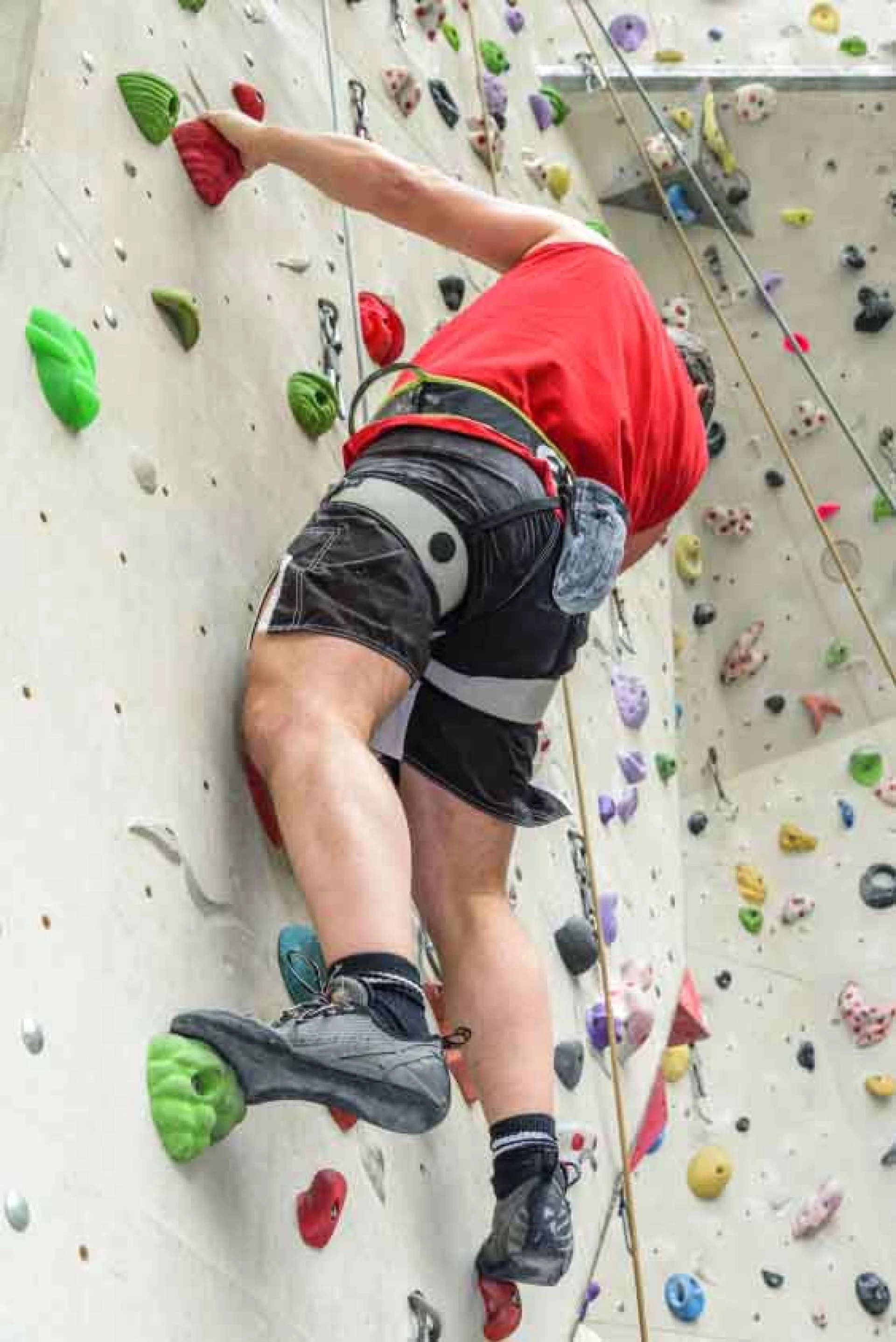 Indoor rock climber ascending a climbing wall with colorful holds, wearing a safety harness and secured by a rope.