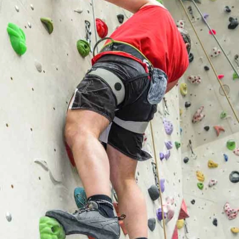 Indoor rock climber ascending a climbing wall with colorful holds, wearing a safety harness and secured by a rope.