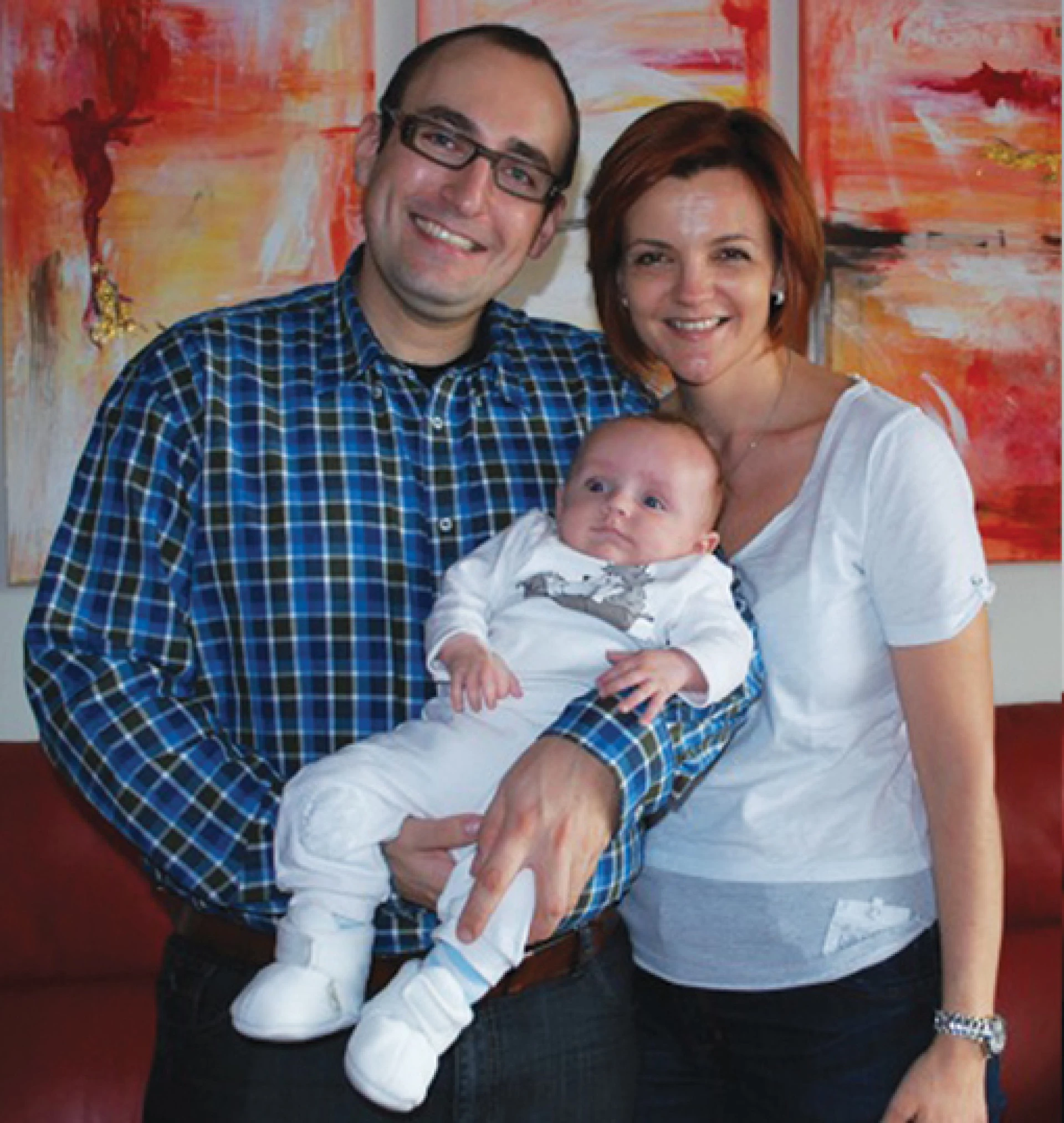 Smiling couple holding a baby indoors with colorful artwork in the background.