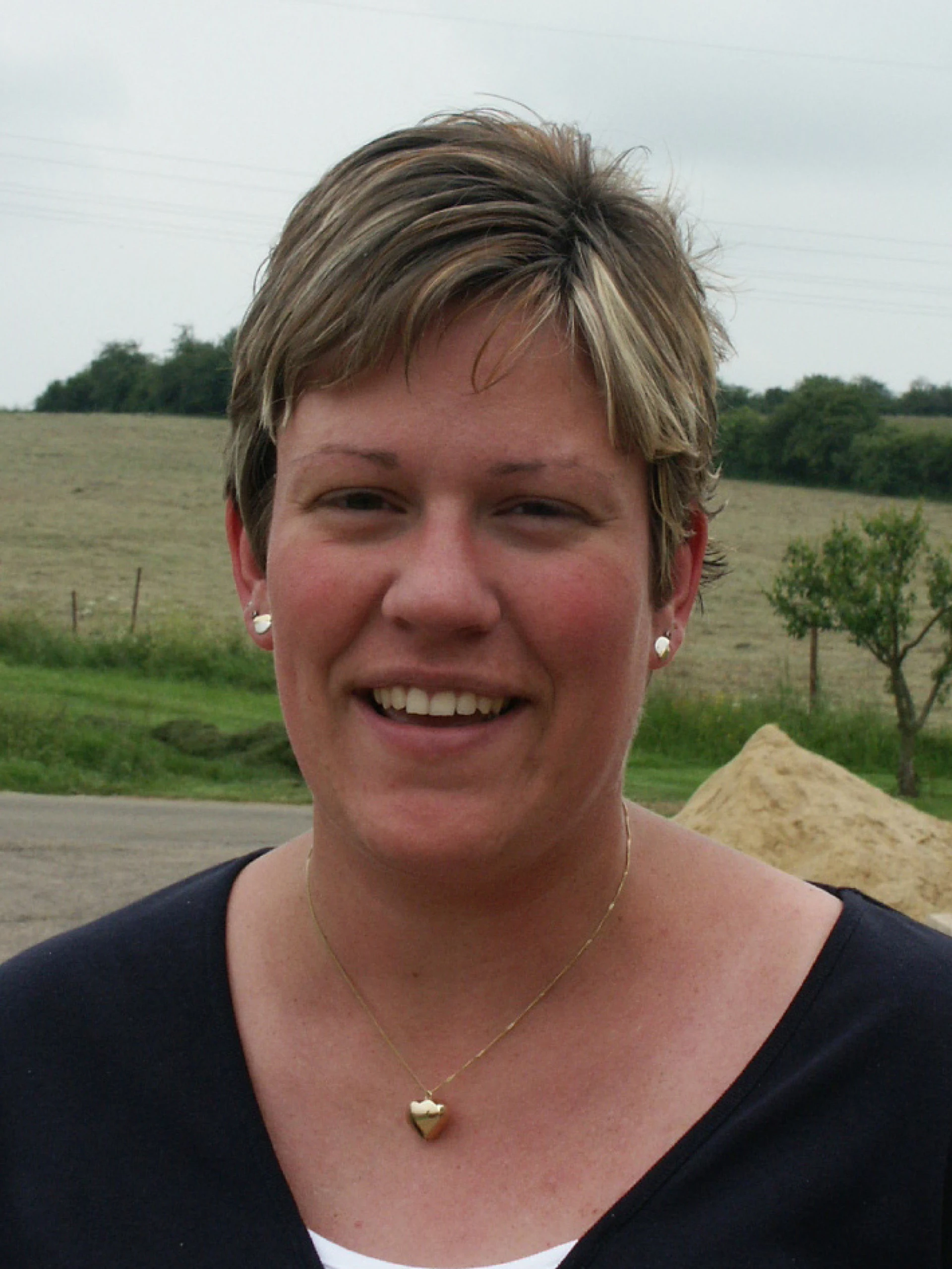 Smiling woman outdoors in a rural landscape with fields in the background.