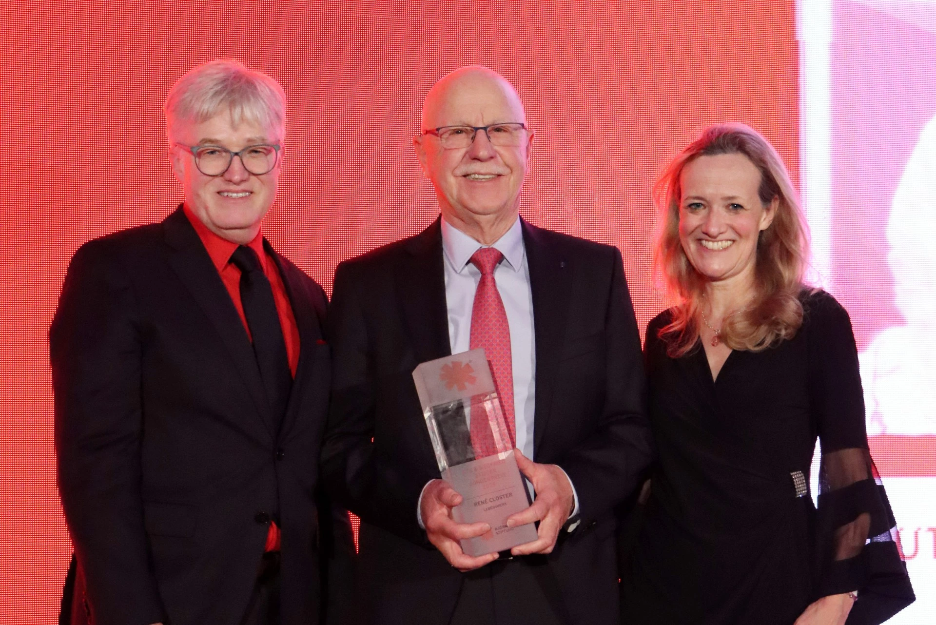 Three people dressed in formal attire pose on stage in front of a red background, with the person in the center holding a glass award. All are smiling, suggesting an award ceremony or formal event.