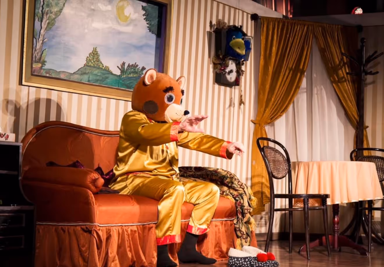Aktor w kostiumie misia siedzący na kanapie, grający swoją rolę na tle scenografii pokoju.