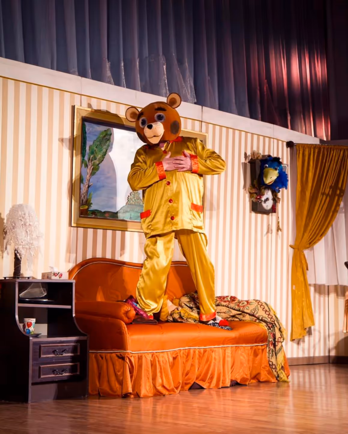 Aktor w kostiumie misia stojący na kanapie, grający swoją rolę na tle scenografii pokoju.