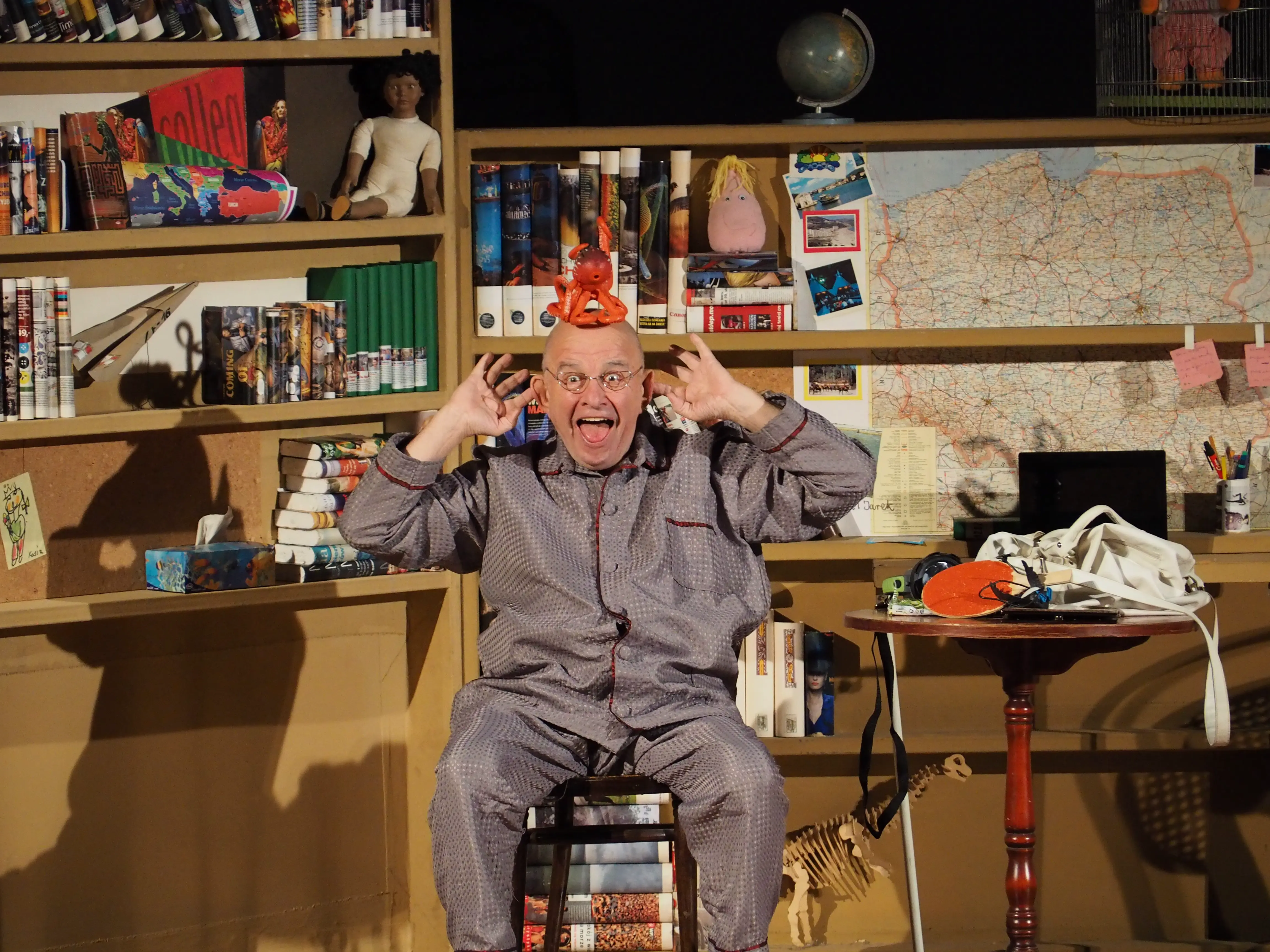 Aktor który robi śmieszną minę, na tle różnych rekwizytów teatralnych.