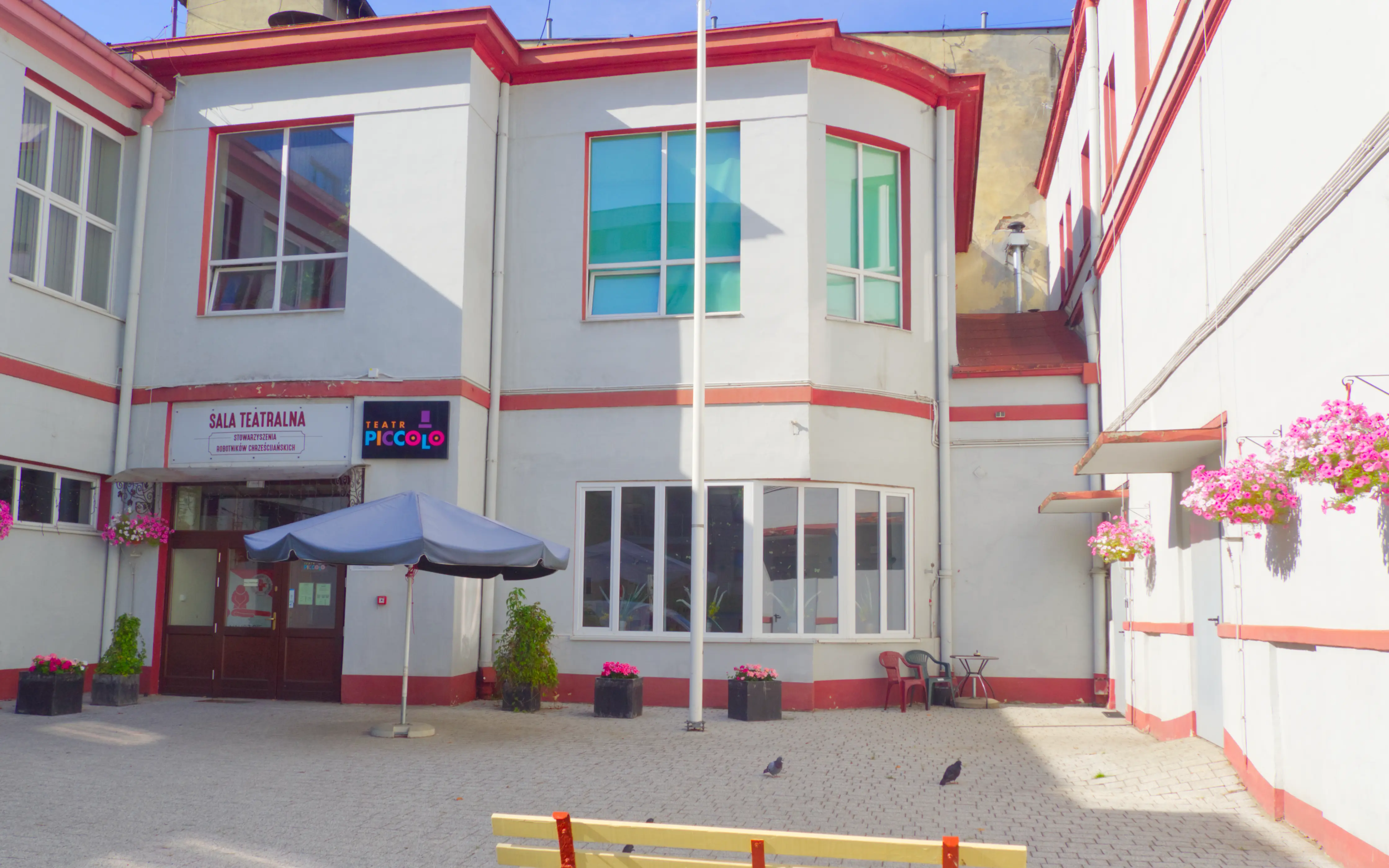 Budynek teatru z zewnątrz – Zdjęcie przedstawiająca fasadę dwupiętrowego budynku z czerwonym dachem i dużymi oknami.