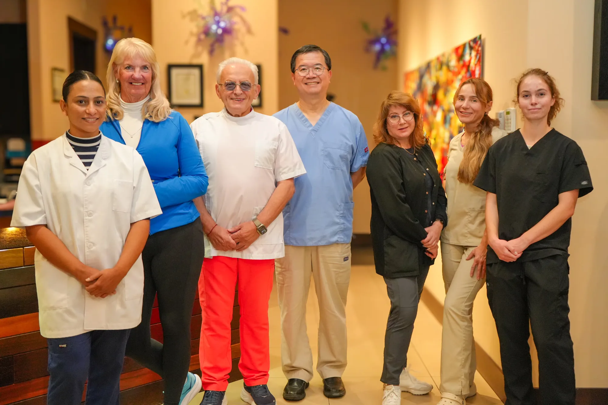 Seven diverse dental professionals standing side by side in a warmly lit clinic hallway, smiling at the camera.