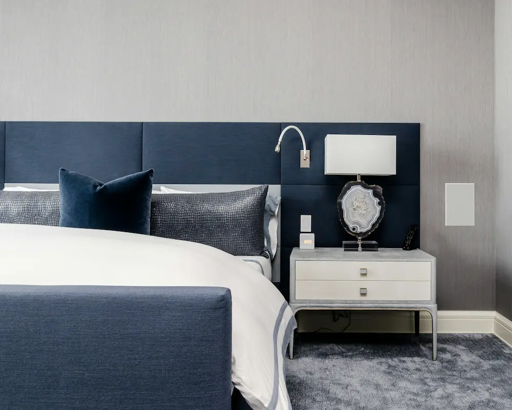 Chambre d'hôtel moderne avec un élégant décor bleu et blanc, un grand lit confortable, des oreillers élégants, une table de chevet avec lampe et une lampe de lecture murale.