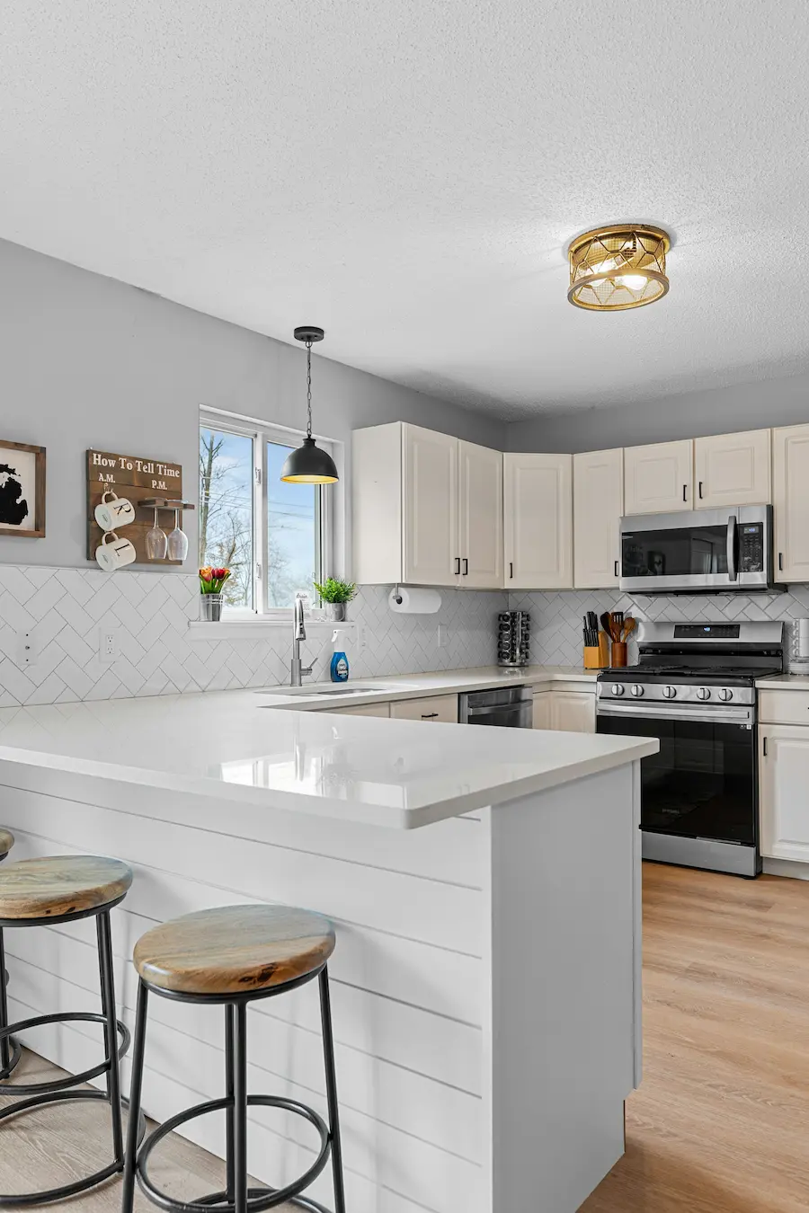 Modern white kitchen with a central island, wooden stools, stainless steel appliances, and bright natural light coming through a window.