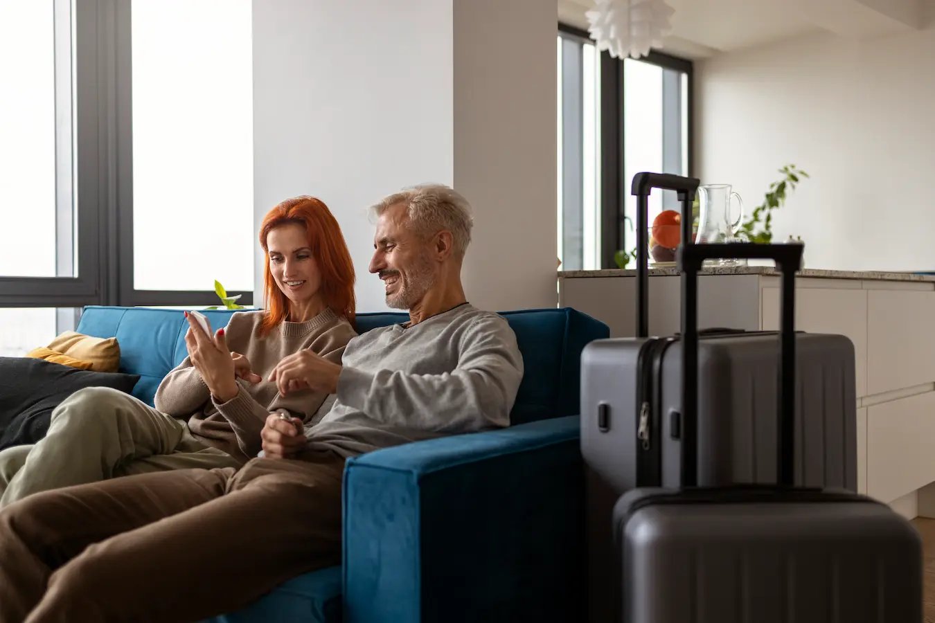 Couple de voyageurs assis sur un canapé dans un appartement moderne, consultant les informations de leur séjour sur un smartphone, avec leurs valises posées à côté.