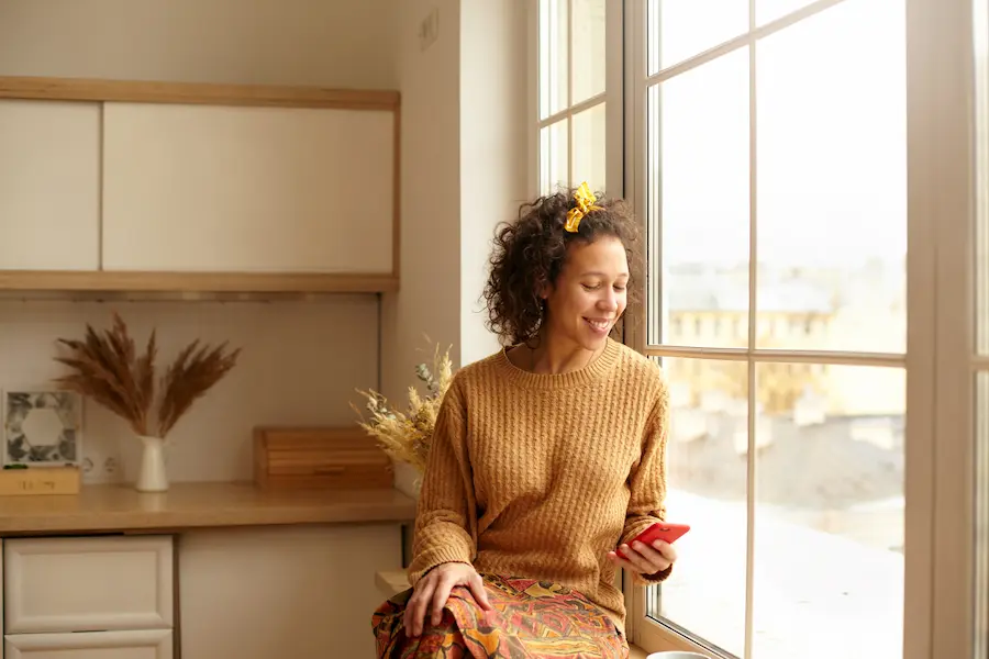 Young woman sitting by a window, smiling while viewing a digital welcome guide on her smartphone in a bright apartment.